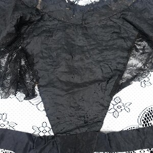 K&ouml;nnte beinhalten: Ein schwarzes Kleidungsst&uuml;ck mit Spitzendetails. Der Stoff scheint ein dunkles, m&ouml;glicherweise seidiges Material zu sein. Das Kleidungsst&uuml;ck hat eine taillierte Form mit Spitzenakzenten an den R&auml;ndern und &Auml;rmeln. Der Hintergrund ist ein wei&szlig;es Spitzemuster.