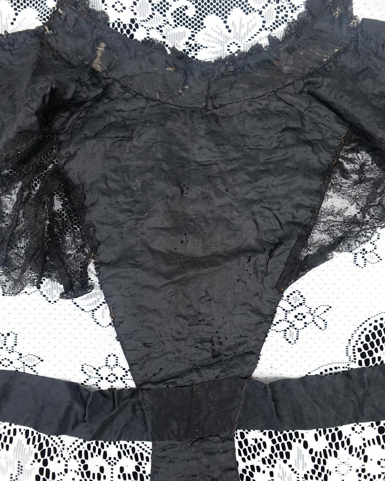 K&ouml;nnte beinhalten: Ein schwarzes Kleidungsst&uuml;ck mit Spitzendetails. Der Stoff scheint ein dunkles, m&ouml;glicherweise seidiges Material zu sein. Das Kleidungsst&uuml;ck hat eine taillierte Form mit Spitzenakzenten an den R&auml;ndern und &Auml;rmeln. Der Hintergrund ist ein wei&szlig;es Spitzemuster.