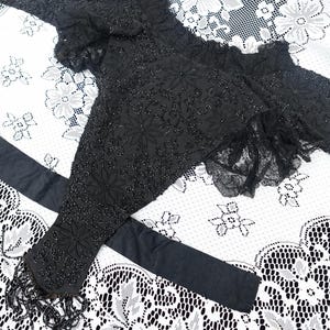 K&ouml;nnte beinhalten: Schwarze, mit Perlen bestickte Bolerojacke mit floralen Stickereien und Fransen. Die Jacke hat lange &Auml;rmel und einen ger&uuml;schten Ausschnitt. Ein schwarzer Sch&auml;rpe ist ebenfalls enthalten. Das Kleidungsst&uuml;ck wird auf einer wei&szlig;en Spitzentischdecke pr&auml;sentiert.