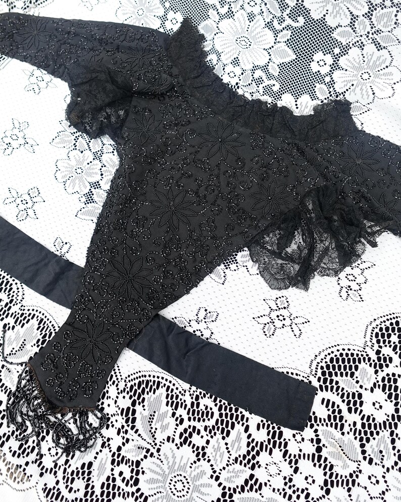 K&ouml;nnte beinhalten: Schwarze, mit Perlen bestickte Bolerojacke mit floralen Stickereien und Fransen. Die Jacke hat lange &Auml;rmel und einen ger&uuml;schten Ausschnitt. Ein schwarzer Sch&auml;rpe ist ebenfalls enthalten. Das Kleidungsst&uuml;ck wird auf einer wei&szlig;en Spitzentischdecke pr&auml;sentiert.