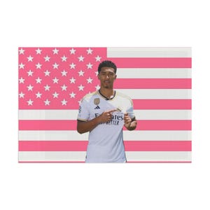 Könnte beinhalten: Ein weißes Fußballtrikot mit schwarzen Akzenten und dem Schriftzug Emirates Fly Better, getragen von einer Person, die auf das Vereinswappen zeigt. Der Hintergrund zeigt ein US-Flaggenmuster mit abwechselnden rosa und weißen Streifen.