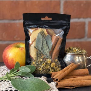 May include: A black resealable pouch containing a blend of dried chamomile flowers, sage leaves, cinnamon sticks, and dried apple slices. The clear front window displays the contents, while the pouch is filled with these aromatic botanical ingredients.