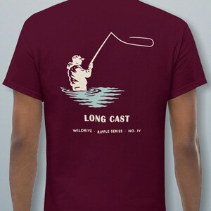 Può includere: Una t-shirt a maniche corte color bordeaux con una grafica color crema e azzurro che raffigura una persona che pratica la pesca a mosca in acqua. Il testo LONG CAST, WILDDRIVE, RIFFLE SERIES e NO. IV è stampato in crema sotto l'illustrazione.