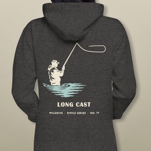 Könnte beinhalten: Ein anthrazitfarbener Kapuzenpullover mit einer Grafik auf der Rückseite, die eine Person beim Fliegenfischen im Wasser zeigt. Das Design enthält den Text "LONG CAST" über "WILDDRIVE . RIFFLE SERIES . NO. IV" in weißer Schrift.