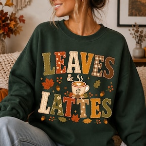 May include: A dark green crewneck sweatshirt featuring a graphic print with the text LEAVES & LATTES. The letters are filled with various autumn-themed patterns including plaid, cable knit, and polka dots, surrounded by maple leaves, acorns, and a latte.