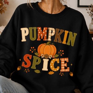 Könnte beinhalten: Ein schwarzes Sweatshirt mit Rundhalsausschnitt und dem Schriftzug PUMPKIN SPICE in großen, gemusterten Buchstaben. Zwischen den Wörtern befindet sich eine orangefarbene Kürbisillustration mit einem braunen Knopf, umgeben von Beeren und Blättern.