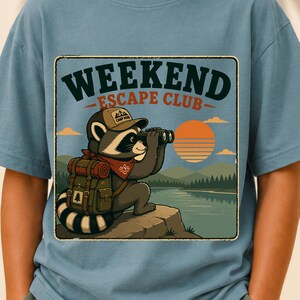 May include: A dusty blue t-shirt featuring a graphic of a raccoon wearing a cap and backpack, looking through binoculars at a sunset over a lake. The text reads WEEKEND ESCAPE CLUB. The illustration includes mountains, trees, and a red bandana.