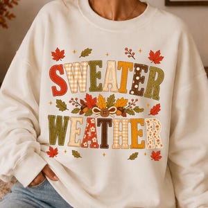 Puede incluir: Una sudadera de cuello redondo color crema con el texto "SWEATER WEATHER" en letras grandes y estampadas. El diseño incluye motivos otoñales como hojas de arce rojas, bellotas y pequeñas bayas en tonos naranja y marrón.