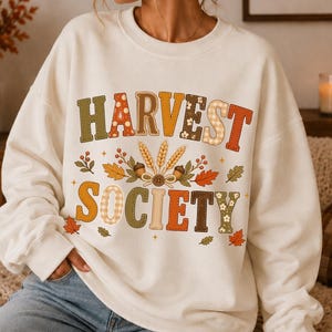 Könnte beinhalten: Ein cremefarbenes Sweatshirt mit Rundhalsausschnitt und dem Schriftzug HARVEST SOCIETY in mehrfarbigen, gemusterten Blockbuchstaben. Zwischen den Wörtern befindet sich eine Grafik aus Weizenähren, Eicheln und Herbstblättern in Orange, Braun und Grün.