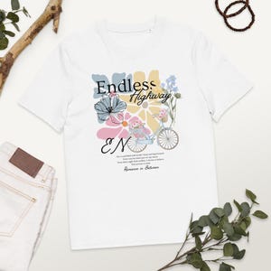 Könnte beinhalten: Weißes T-Shirt mit floralem Design und den Worten "Endless Highway". Das Design umfasst ein hellblaues Fahrrad mit einem Blumenkorb, vor einem Hintergrund aus Pastellblumen. Der Text "EN" ist ebenfalls enthalten.