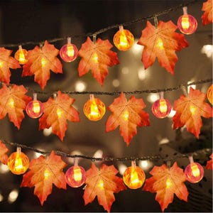 May include: A string of decorative lights featuring alternating artificial maple leaves in shades of orange, red, and yellow, interspersed with small, round, glowing light bulbs that emit a warm, soft illumination along a thin dark wire.