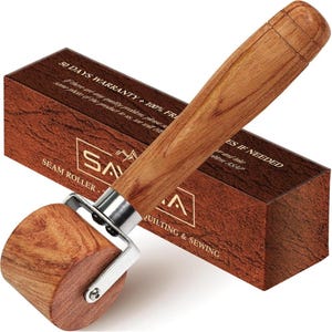 May include: A seam roller with a cylindrical wooden wheel and a long wooden handle connected by a silver metal bracket. The tool is positioned in front of a rectangular brown box featuring the text SAVANA, SEAM ROLLER, and QUILTING & SEWING.