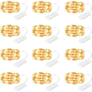 May include: A set of twelve identical copper wire string lights, each coiled into a circular shape and connected to a small, rectangular white battery pack by a short white cord. The lights emit a warm, glowing light throughout the coils.