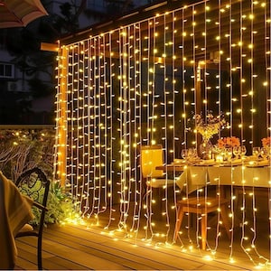 May include: Warm white string lights hanging from a wooden structure, creating a curtain effect. The lights illuminate a dining table set for a meal, with flowers and candles. The scene is set outdoors on a wooden deck, creating a cozy ambiance.