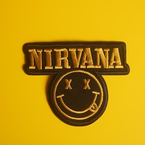 May include: A black and gold Nirvana patch featuring the band's logo and a smiley face with crossed-out eyes and a protruding tongue. The patch has a black border and is set against a yellow background.