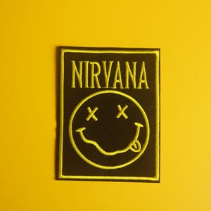 May include: A black patch with the word "NIRVANA" in yellow, above a yellow smiley face with crossed-out eyes and a protruding tongue. The patch has a yellow border and is set against a yellow background.