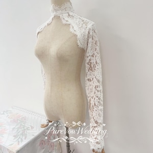 Puede incluir: Un bolero de encaje blanco con cuello alto y mangas largas con bordes festoneados. El delicado patrón floral de encaje cubre los hombros y los brazos, ofreciendo una apariencia transparente. El texto PureVouWedding es visible.