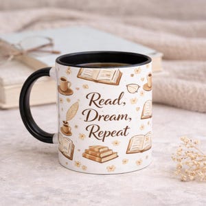 Puede incluir: Taza de cerámica blanca con asa e interior negros, decorada con ilustraciones de libros, tazas de café y flores. La taza tiene el texto "Read, Dream, Repeat" en escritura marrón. La taza está llena de un líquido oscuro.