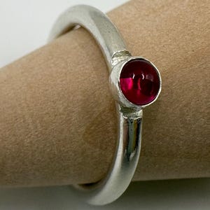 May include: A silver ring with a round, deep red gemstone. The ring has a simple, elegant design with a polished finish. The gemstone is set in a bezel setting, and the band is smooth and rounded.