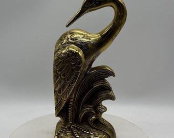 May include: A polished brass figurine of a crane standing among stylised waves. The bird is depicted with its head turned back, showcasing detailed feather textures on its wings and a smooth, curved neck, all set on a textured base.