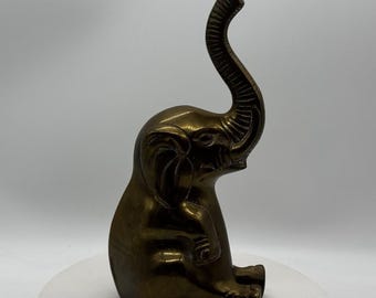 Può includere: Una statuetta di elefante in ottone massiccio in posizione seduta con la proboscide rivolta verso l'alto. La superficie metallica presenta una patina scura e lucida con una trama visibile sulla proboscide e sulle orecchie.