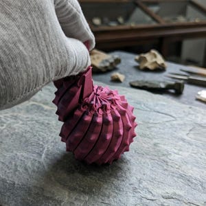 Op de afbeelding: Een magenta 3D-geprint object met een stekelig, bolvormig ontwerp. Het object wordt vastgehouden door een gehandschoende hand. De achtergrond toont een stenen oppervlak en diverse gereedschappen en objecten.