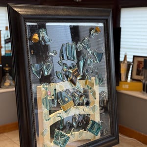 Op de afbeelding: Een ingelijst kunstwerk met fragmenten van heldere en blauwgroene glazen flessen, gerangschikt om een uniek abstract ontwerp te creëren. Het kunstwerk bevat flesetiketten en kurken, allemaal ingesloten in een zwart houten frame. De achtergrond is lichtbeige.