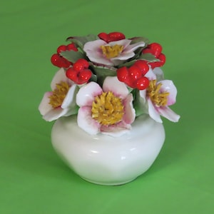 May include: A white ceramic vase with a bouquet of flowers and red berries. The flowers have white petals with pink edges and yellow centers. The arrangement sits on a green background.