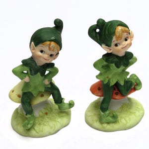May include: Two ceramic figurines of elves. Each elf is wearing a green outfit with a matching hat. One elf is sitting on a mushroom with yellow spots, and the other is standing on a mushroom with red spots. Both are on a green base.