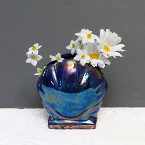 May include: A blue iridescent seashell-shaped vase holding white and yellow daisies. The vase has a square base and a textured surface. The flowers are clustered at the top of the vase.