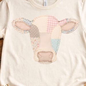 May include: Cream-colored t-shirt featuring a patchwork cow face design. The cow's face is made of various pastel-colored fabrics, including pink, blue, and floral patterns. The shirt is displayed on a wooden surface with a teacup, books, and a straw hat.
