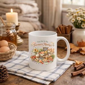 May include: White ceramic mug with a farmer's market design. The mug features a colorful illustration of fresh produce, bread, and sunflowers, with the text "Take Me To The Farmers Market". The mug is on a checkered cloth.