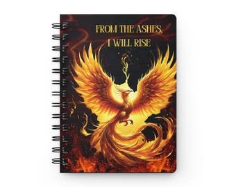 May include: A spiral-bound notebook featuring a black cover with a vibrant orange and yellow phoenix illustration in the center. The text "FROM THE ASHES, I WILL RISE" is printed in gold lettering above the phoenix, set against a fiery background.
