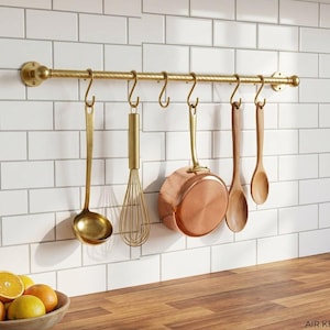 Könnte beinhalten: Eine Küchenutensilienhalterung aus Messing mit hängenden Werkzeugen vor einer weiß gefliesten Wand. Die Halterung hält eine Schöpfkelle, einen Schneebesen, eine Kupferpfanne und Holzlöffel. Eine Schüssel mit Orangen steht auf einer Holztheke.