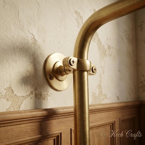 Op de afbeelding: Een messingkleurige gebogen buis bevestigd aan een muur met een cirkelvormige beugel en een klem. De muur heeft een getextureerde, gebroken witte afwerking met bruine houten panelen aan de onderkant. De buis is met schroeven bevestigd.