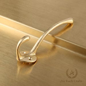 Puede incluir: Un gancho de pared de color latón con un diseño curvo. El gancho tiene un extremo redondeado y una base circular con dos orificios para tornillos. El gancho está sobre una superficie dorada.