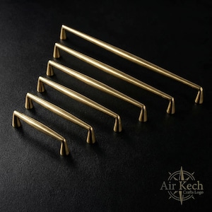 May include: A collection of six brass-colored cabinet pulls arranged diagonally on a black surface. The pulls vary in length, with a cylindrical center and angled ends. The Air Kech Crafts logo is in the bottom right corner.
