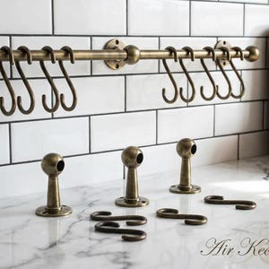 Może przedstawiać: Antyczny mosiężny reling kuchenny z hakami S, wspornikami montażowymi i dodatkowym osprzętem. Szyna jest zamontowana na białej, kafelkowej ścianie. Dodatkowy sprzęt obejmuje trzy dystanse i haki S.