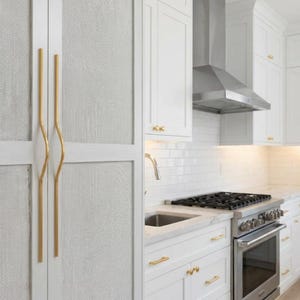 Può includere: Una cucina bianca con mobili bianchi, un fornello in acciaio inossidabile e una cappa in acciaio inossidabile. Le ante dei mobili hanno maniglie color oro. Le ante dei mobili hanno un pannello strutturato tessuto.