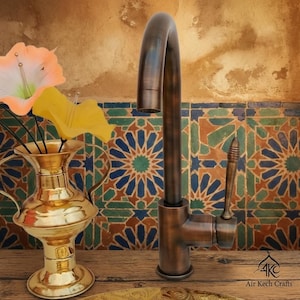 Op de afbeelding: Een bronzen keukenkraan met een gebogen uitloop en hendel. De kraan staat tegen een achtergrond van tegels met patronen in blauw, groen en oranje. Een gouden vaas met bloemen staat op de voorgrond.