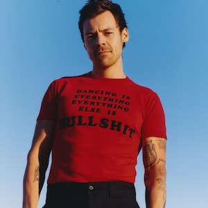 Könnte beinhalten: Ein rotes Kurzarm-T-Shirt mit schwarzem Text, der DANCING IS EVERYTHING EVERYTHING ELSE IS BULLSHIT lautet. Das Shirt hat einen Rundhalsausschnitt und wird mit einer schwarzen Hose getragen. Der Schriftzug ist mittig auf der Brust platziert.