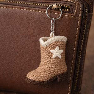 Op de afbeelding: Een gehaakte sleutelhanger in de vorm van een cowboylaars. De laars is bruin met een witte ster en een crèmekleurige rand. Hij hangt aan een zilverkleurige ring en ketting. De achtergrond is een bruine leren portemonnee. De laars is ongeveer 7 cm hoog.