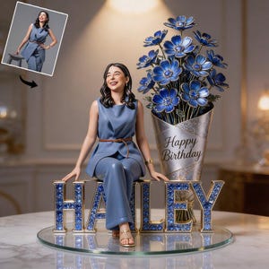 May include: A blue figurine of a person sitting on a bedazzled name, with a vase of blue flowers and the words "Happy Birthday". The figurine is wearing a blue outfit and a brown belt. The name is "HAILEY".