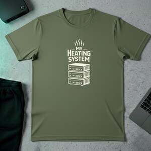 Op de afbeelding: Olijfgroen t-shirt met de tekst "MY HEATING SYSTEM" boven een witte afbeelding van drie gestapelde elektronische apparaten. Het t-shirt ligt op een lichtgrijze ondergrond.