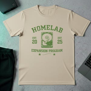 Peut inclure: T-shirt beige avec un motif vert. Le motif comprend le mot "HOMELAB" au-dessus d'une illustration de disque dur. En dessous, les mots "EST. 20" et "EST. 25". Les mots "EXPANSION PROGRAM" sont en dessous du disque dur.