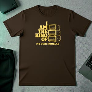 Op de afbeelding: Een bruin t-shirt met de tekst "I AM THE KING OF MY OWN HOMELAB" in het geel. De afbeelding toont een stapel van drie gele elektronische apparaten. Het shirt met korte mouwen is gemaakt van zacht materiaal.