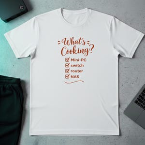 Puede incluir: Camiseta blanca con la frase "What's Cooking?" en marrón. Debajo, casillas de verificación junto a las palabras "Mini-PC", "switch", "router" y "NAS". La camiseta está hecha de un material suave.