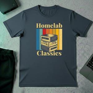 May include: Dark navy blue t-shirt with the text "Homelab Classics" in a retro style. The design features a stack of three server-like devices with a colourful striped background. The shirt is made of a soft material.