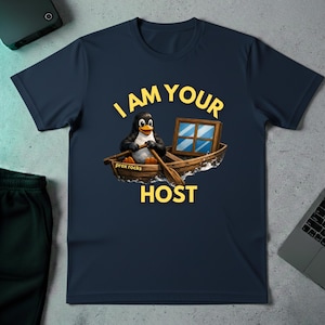 Op de afbeelding: Marineblauw t-shirt met een afbeelding van een pinguïn in een boot, met roeispanen en een raam. De tekst luidt "I AM YOUR HOST" in geel, met "prox rocks" eronder. Een casual t-shirt.