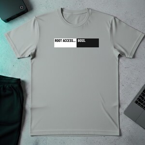 May include: A light gray t-shirt with the text "ROOT ACCESS... BOSS." in a white and black rectangular design across the chest. The shirt is short-sleeved and appears to be made of a soft material. A pair of dark green shorts and a laptop are also visible.